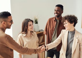 Four colleagues smiling and shaking hands in a bright office setting.